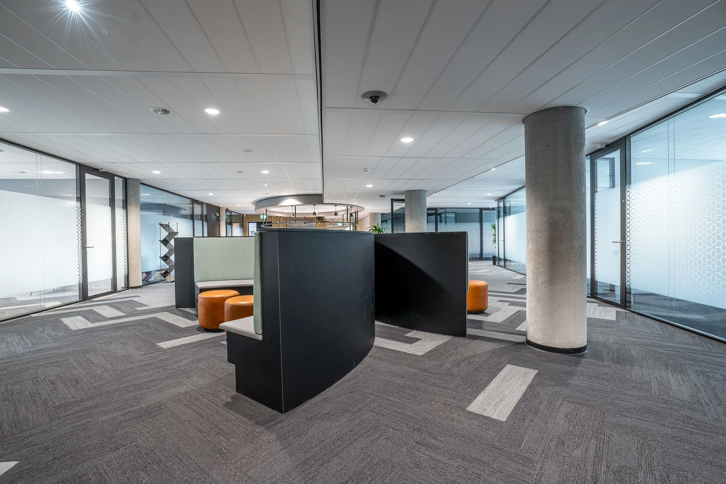 Empty office floor with booth seating, concrete pillars, glass-walled meeting rooms and a suspended ceiling